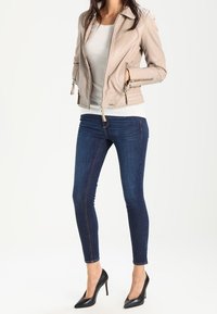 Chaqueta de moto de cuero beige con puños con cremallera, combinada con jeans ajustados de color azul oscuro y tacones negros de punta. Top de color sólido debajo.