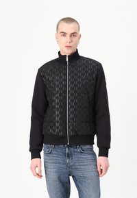 Svart jacka som kombinerar vadderad framdel med geometriskt mönster och tröjliknande ärmar. Har hög krage och framzipper.