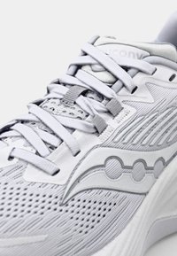 Baskets de sport gris clair avec tige en mesh, col rembourré et lacets plats. Présente un design texturé et un logo près du talon, mettant en avant la respirabilité.