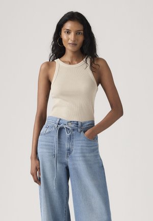 Kvinde med langt krøllet hår iført beige ærmeløs top og løse lyseblå jeans med snørebælte, poserer med den ene hånd i lommen.