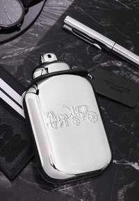 Flacone in metallo argento con una superficie liscia e riflettente, caratterizzato da un design in rilievo di un cavallo e una carrozza. Include un'etichetta nera con la scritta "Coach New York."