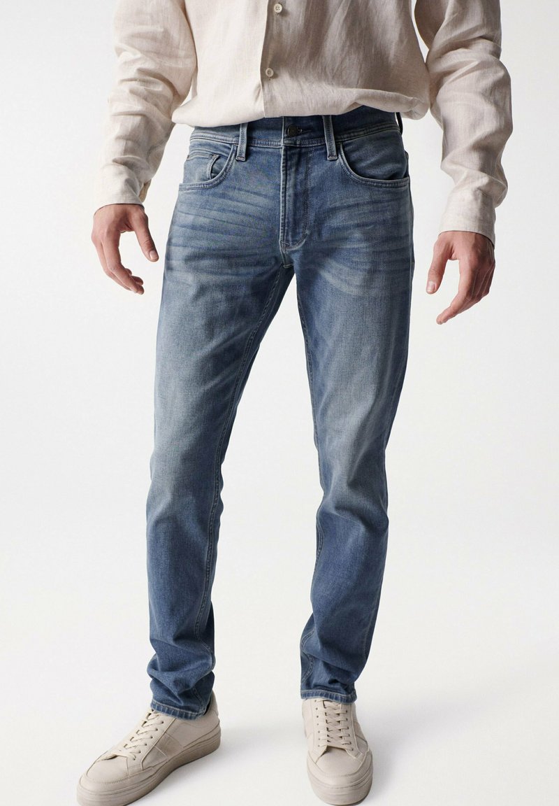 Jeans de mezclilla azul claro con corte slim, textura ligeramente desgastada y diseño clásico de cinco bolsillos, combinados con zapatillas beige y una camisa clara.