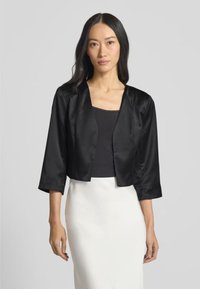 Giacca nera in satin corta con maniche tre quarti, con scollatura rotonda e texture liscia, indossata sopra un top nero e una gonna bianca.