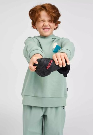 Jong roodharig kind in muntgroene outfit die een zwarte Reima-hoed naar voren rekt met gesloten ogen en een glimlach.