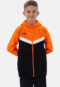Schwarze und orange Zip-Jacke mit Kapuze, ausgestattet mit einem weißen Chevron-Design, strukturiertem Stoff und seitlichen Reißverschlusstaschen. "JAKO"-Logo auf der Vorderseite.
