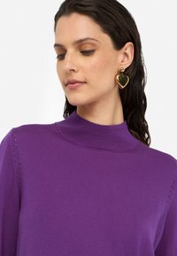Maglione a collo alto di lana viola, con texture a coste e dettagli a taglio sulle spalle. Indossato con orecchini a forma di cuore dorati, con finitura lucida.