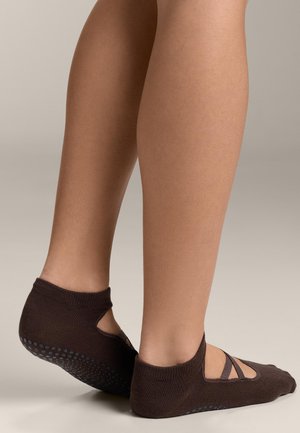 Picioare purtând șosete maro închis cu talpă antiderapantă și curele încrucișate, ridicate pe vârfuri, pe un fundal neutru.