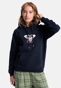 Sweat-shirt à capuche bleu marine avec un grand logo en forme de cœur et de diamant, présentant des accents blancs et roses, un cordon de serrage ajustable et une poche avant.