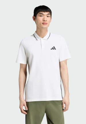 Młody mężczyzna stojący, ubrany w białą koszulkę polo Adidas z czarnym logo i zielone spodnie na jednolitym jasnym tle.