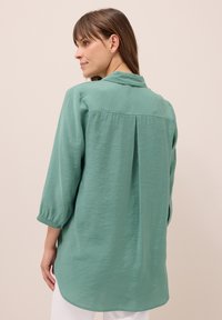 Chemise vert clair à manches longues avec poignet à boutons, pli au dos et col. Tissu doux et texturé avec une coupe décontractée.
