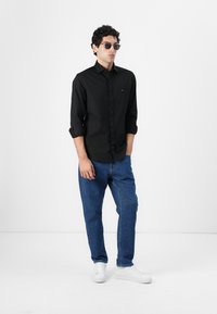 Svart skjorte med knapper og rullede ermer, blå jeans og hvite sneakers. Tilbehør inkluderer grå solbriller. Enkelt design og klassisk passform.