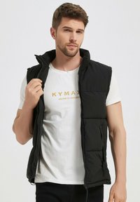 Gilet matelassé noir avec une fermeture éclair à l'avant, un col montant et des poches latérales. Porté sur un t-shirt blanc avec un texte doré. Texture lisse.