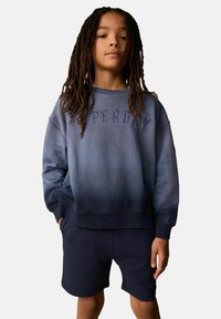 Felpa con gradiente blu e logo "SUPERDRY" in rilievo, abbinata a pantaloni corti scuri. Tessuto in cotone, vestibilità rilassata, polsini e orlo a coste.