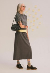 Polo shirt in maglia grigia, top a strati giallo, gonna marrone alla caviglia, borsa nera, scarpe marroni con lacci, braccia tatuate, sfondo neutro.