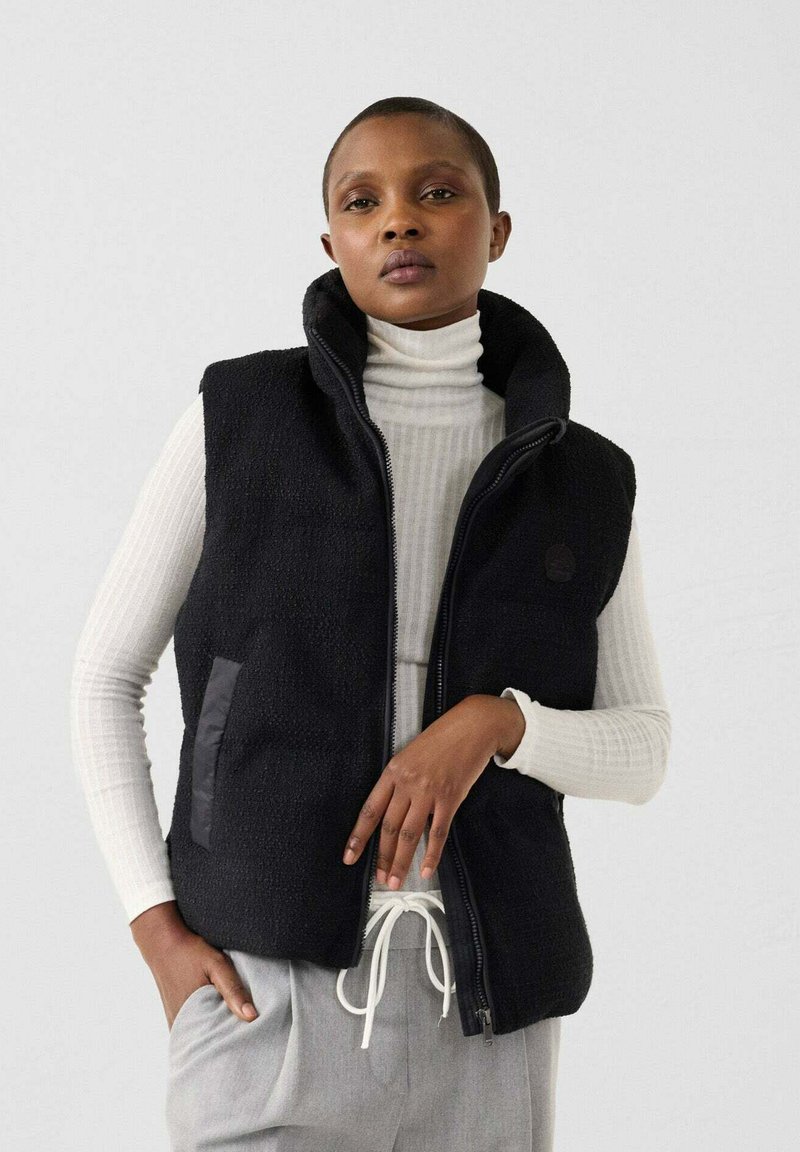 Donna che indossa un gilet in pile nero con cerniera sopra un dolcevita a costine bianco con pantaloni grigi chiaro con coulisse, in piedi davanti a uno sfondo bianco neutro.