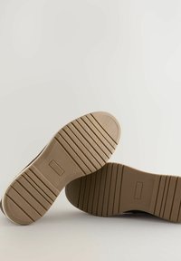 Suelas de dos zapatos marrones con surcos horizontales y patrones texturizados sobre un fondo claro y liso.