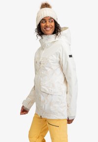 Witte geïsoleerde jack met een gestructureerd patroon, verstelbare capuchon en twee voorzakken, gecombineerd met felgele broek en een beige beanie.