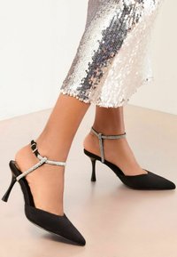Pantofi cu toc slingback negri, cu vârful ascuțit și toc stiletto. Un brâu decorativ cu strasuri la gleznă adaugă detalii. Superior dintr-un material textil neted, cu un finisaj elegant.