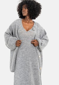 Jasnoszary, oversize kardigan wykonany z dzianiny z dużymi rękawami, ozdobiony okrągłymi guzikami i teksturowanymi akcentami. Noszony na dopasowaną sukienkę.