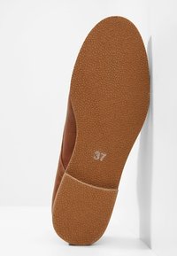 Semelle de chaussure marron avec un grip en caoutchouc texturé et le numéro de taille 37 imprimé près du centre, inclinée contre un mur blanc.