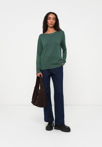Green knitted sweater with a wide neckline, paired with dark blue flared jeans. Model holds a brown suede bag and wears black platform shoes.