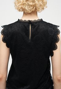 Άτομο με κοντά σγουρά ξανθά μαλλιά που φοράει μαύρο μπλουζάκι από δαντέλα με μανίκια με κυματιστές άκρες και κλείσιμο με κουμπί σε σχήμα κλειδαρότρυπας στον πίσω λαιμό.