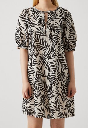 Femme portant une robe beige mi-longue avec imprimé feuilles noires, manches courtes bouffantes et un décolleté goutte d'eau noué devant avec des nœuds.