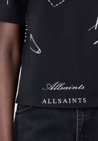 Musta musta t-paita, jossa on valkoisia graafisia kuvastoja, kuten turvapitimiä ja tekstiä. Helma esittelee "Allsaints"-logon kahdessa tyyliissä.