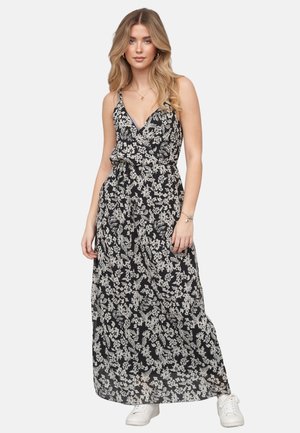 Młoda kobieta z długimi blond włosami nosi czarno-białą sukienkę maxi w kwiaty oraz białe trampki, stoi na jednolitym tle.