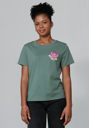 T-shirt en coton vert à col rond, arborant un graphique de fleur de lotus rose sur la partie gauche de la poitrine. Manches courtes et coupe décontractée.
