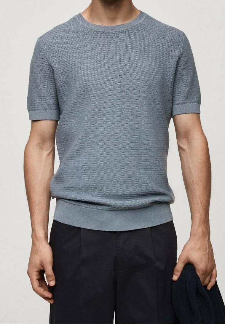 Homme portant un pull en maille texturée bleu clair à manches courtes et un pantalon foncé, tenant un vêtement foncé dans sa main droite.