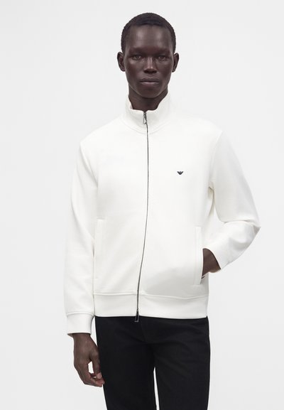 Homme portant une veste blanche zippée avec des poches latérales et un pantalon noir, debout devant un fond clair uni.