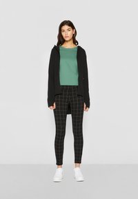 Un sweat à capuche noir zippé sur un haut vert, associé à des leggings à carreaux noirs et des baskets blanches. Tissu lisse, design ajusté et couleurs contrastantes.