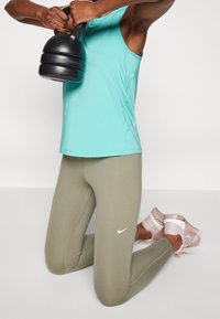 Czarna kettlebell trzymana obiema rękami; na sobie turkusowy top na ramiączkach, oliwkowe legginsy i jasnoróżowe buty sportowe z siateczkowymi detalami.