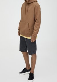 Sudadera marrón con bolsillo canguro, llevada sobre una camiseta amarilla. Combinada con pantalones cortos de mezclilla negros y zapatillas negras. Fondo liso.