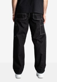 Pantaloni di jeans neri con gamba larga, cuciture visibili, molteplici tasche e una texture che suggerisce durata. Indossati con sneakers bianche.