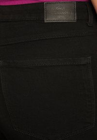 Pantalones de mezclilla negros con una textura suave, que cuentan con un bolsillo trasero y una etiqueta de cuero grabada con "ONLY Garmakoma Denim."