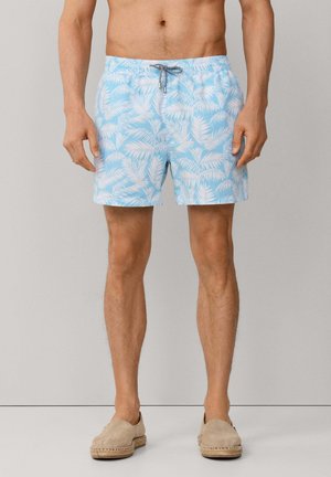 Hombre con pantalones cortos de baño azul claro con estampado blanco de hojas de palmera y alpargatas beige sin cordones, de pie frente a un fondo liso.