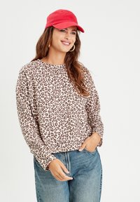 Top a maniche lunghe con stampa leopardata in beige e marrone, tessuto morbido, scollatura rotonda, vestibilità rilassata, abbinato a jeans blu e un cappellino rosso.
