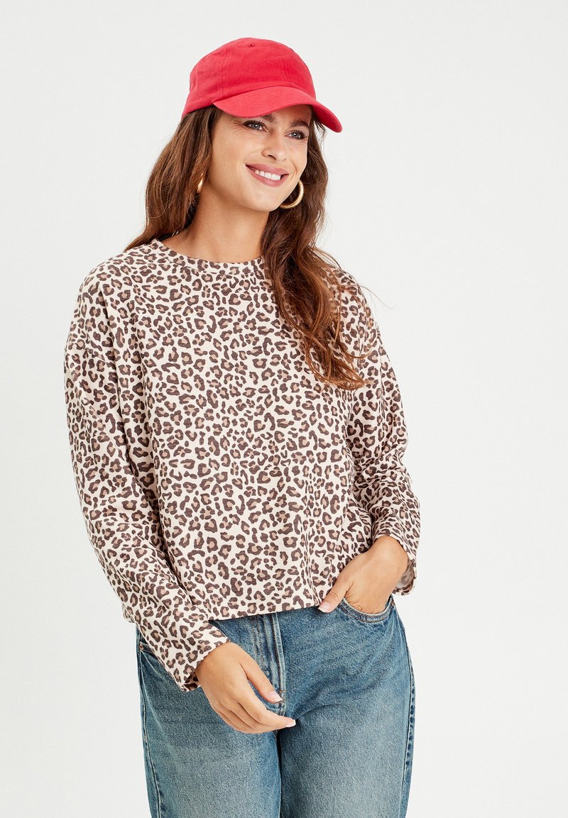 Top a maniche lunghe con stampa leopardata in beige e marrone, tessuto morbido, scollatura rotonda, vestibilità rilassata, abbinato a jeans blu e un cappellino rosso.
