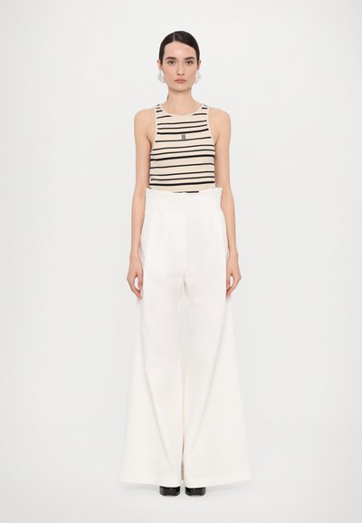 Top senza maniche a strisce nere e beige abbinato a pantaloni bianchi a gamba larga con vita alta. Modella che indossa stivali neri alla caviglia.