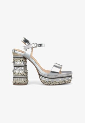 Silver platform sandals with a chunky embellished heel featuring intricate textures, metallic accents, and a glossy finish.