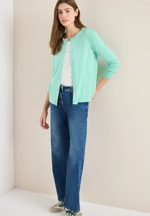 Ung kvinde står op ad en væg iført en mintgrøn cardigan med knapper, hvid top, blå jeans og beige sneakers med grønne detaljer.