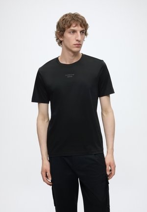 Jeune homme aux cheveux bouclés portant un t-shirt noir Porsche BOSS et un pantalon noir, debout devant un fond blanc uni.
