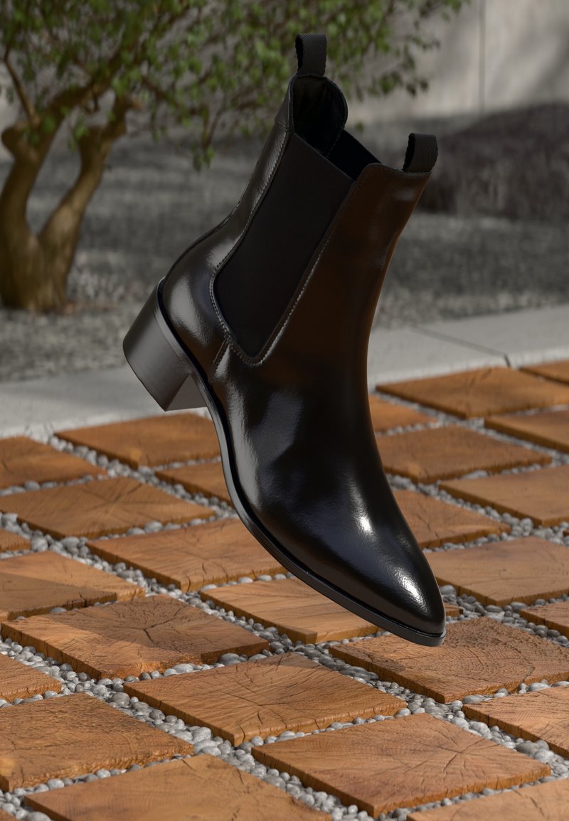 Bota Chelsea em couro preto com ponta afilada, painéis laterais elásticos e um salto robusto, descansando sobre uma superfície com padrão de pedra e madeira.