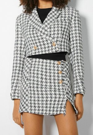 Femme portant un blazer court et une jupe assortie à motif pied-de-poule noir et blanc, avec des boutons dorés et une fente à l'avant.