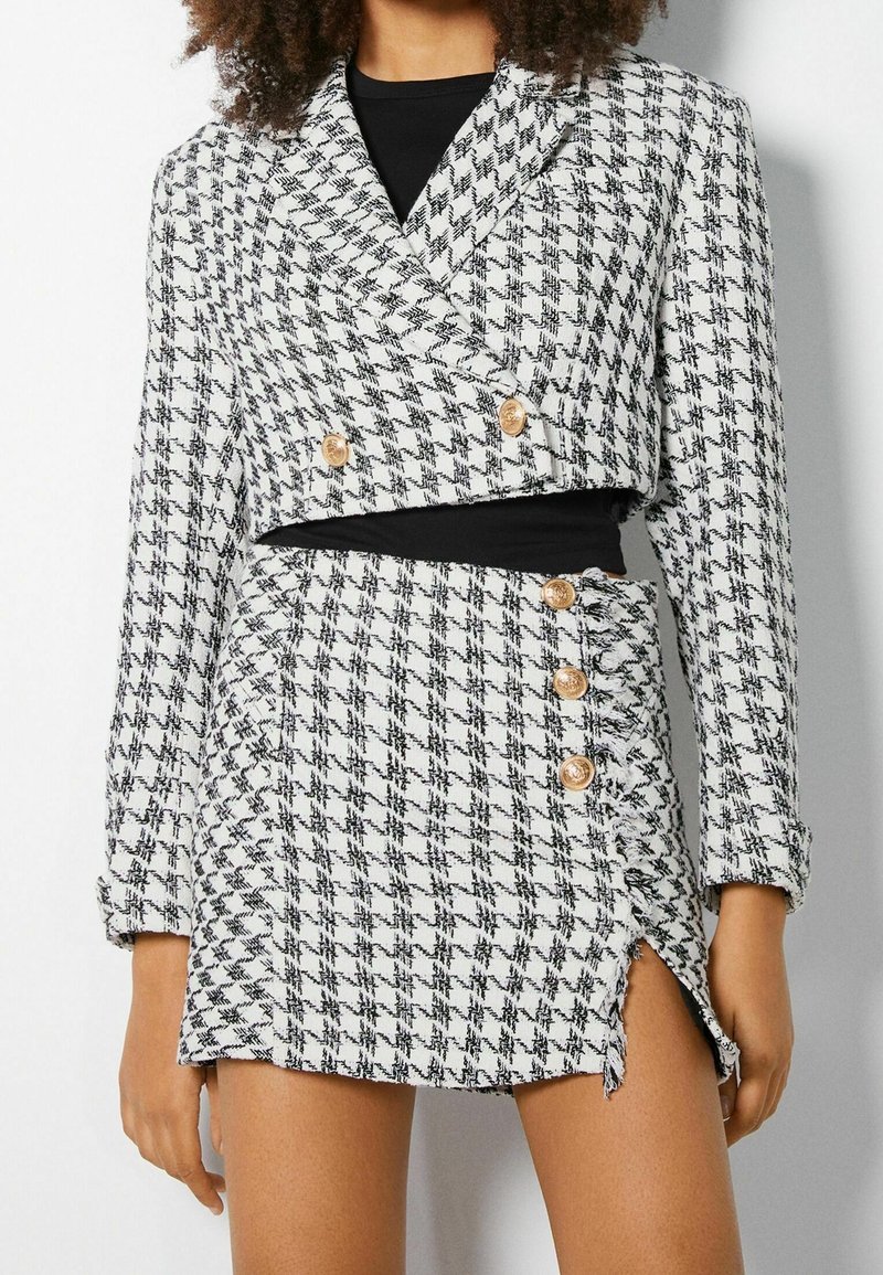 Femme portant un blazer court et une jupe assortie à motif pied-de-poule noir et blanc, avec des boutons dorés et une fente à l'avant.