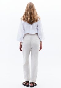 Vrouw met lang haar staat met haar gezicht naar weg, gekleed in een witte blouse, lichtbeige broek en donkerbruine sandalen op een witte achtergrond.