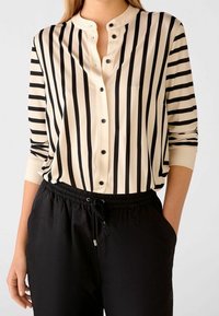 Proužkovaná halenka v krémové a černé barvě, bez límce, s knoflíkovou légou a dlouhými rukávy. Kombinována s černými kalhotami na šňůrku.