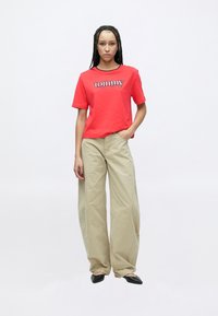 Raudonas medvilninis marškinėlis su „tommy“ logotipu, derinamas su kreminės spalvos kelnėmis plačiais kojų siūlais. Modelis dėvi juodus smailius batus. Vienodas baltas fonas.